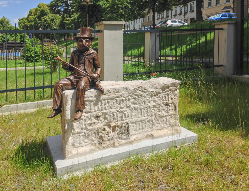 Bronzeplastik des Museumsmaskottchens, Kater Franz Adelbert Hüthling. Typisch gekleidet im Anzug mit Spazierstock und Zylinder, hier sitzend auf einem steinernen Sockel im Freien vor einem Zaun im Museumgarten.