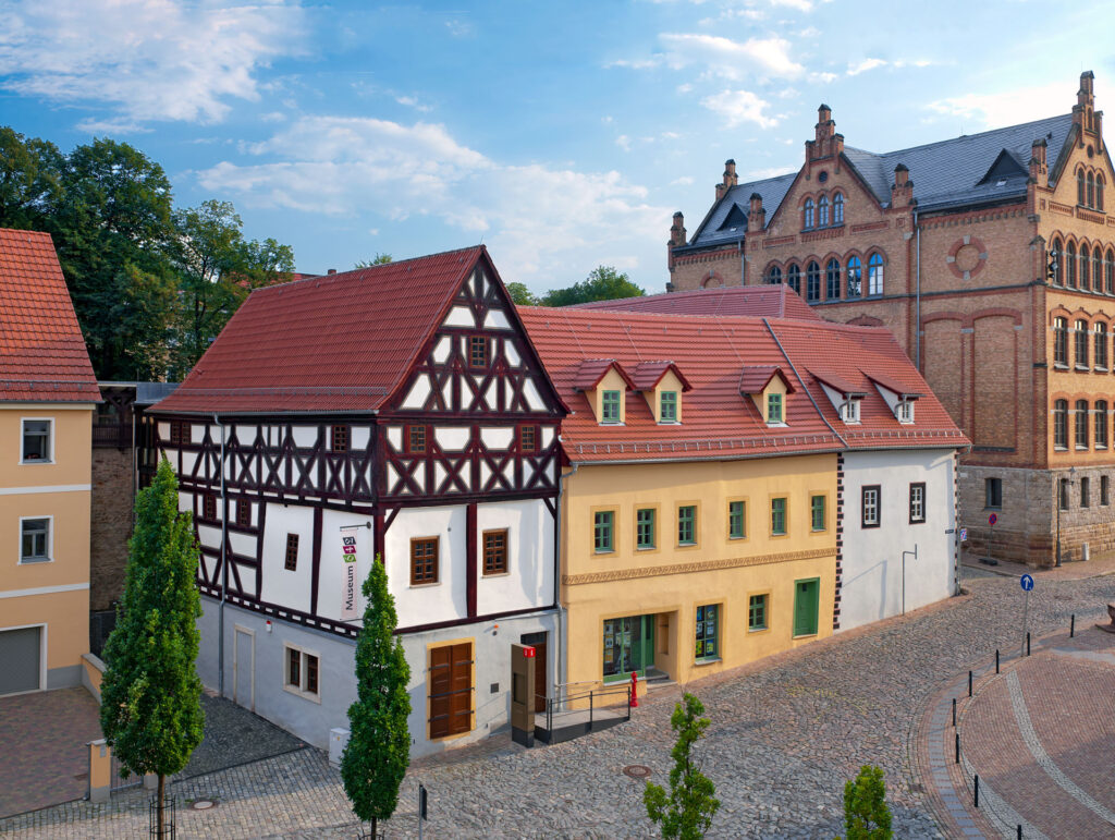 Das Museumsensembles aus drei historischen Gebäuden mit roten Dachziegeln am gepflasterten Klosterplatz 2-4-6. Haus 6 links mit Fachwerk und weißem Anstrich sowie einem gelben Anstrich am mittleren Haus 4 und einem Weißen an Haus 2. Am rechten Bildrand neben Haus 2 steht ein großes, gelbes Backsteingebäude, das Pößnecker Gymnasium „Am Weißen Turm“.
