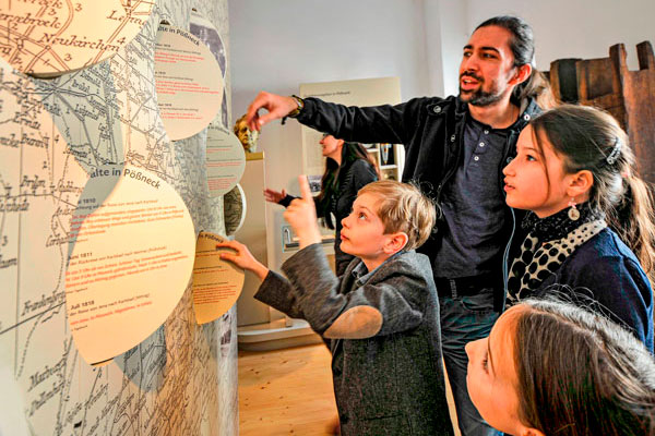 Ein Mann und drei Kinder stehen vor einer Ausstellungswand mit einer historischen Karte der früheren Postwege und bewegen die interaktiven Drehscheiben. Darauf sind Tagebucheinträge Johann Wolfgang von Goethes von seinen Reisen zu finden.