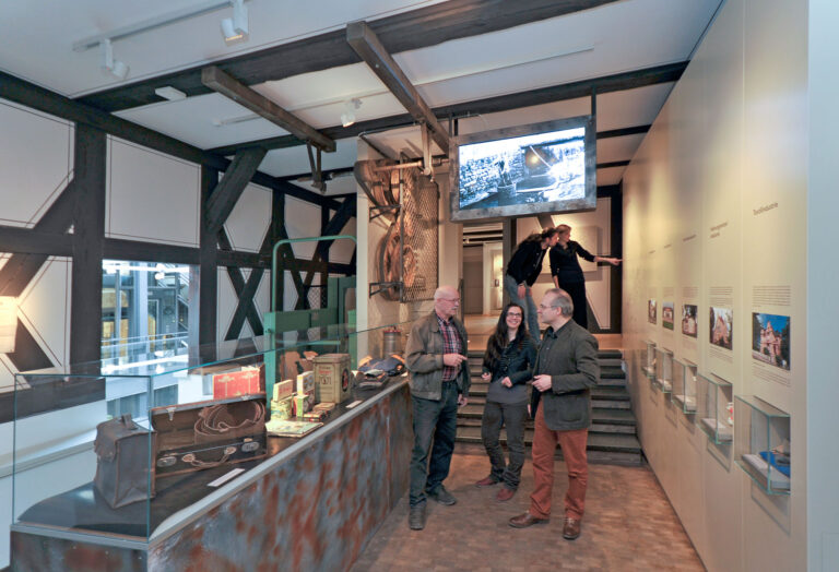 Fünf Personen stehen im Ausstellungsraum zur Industrialisierung in Pößneck. An der rechten Seite eine lange Ausstellungswand mit Texten zu den Pößnecker Fabrikantenvillen der Gründerzeit und historischen Objekten in kleinen Vitrinen. Der Stadtplan auf der Tafel im Hintergrund veranschaulicht die in Pößneck vertretenen Industriezweige und Firmen. Einblicke in den Fabrikalltag des 19. Jahrhunderts vermittelt der Monitor, welcher von der Decke hängt und mit dem damals genutzten Transmissionsantrieb links verbunden ist. Auf der Nachbildung eines Förderbandes in der Mitte des Raumes sind Industrieerzeugnisse aus Pößneck ausgestellt.