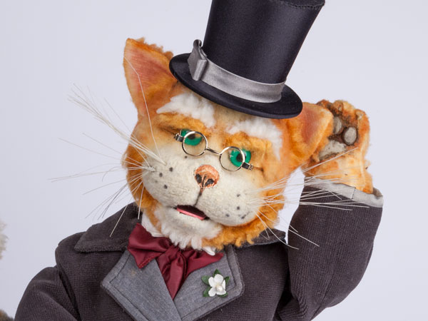 Museumsmaskottchen Franz Adelbert Hüthling ist eine Katerpuppe mit orangefarbenem Fell und trägt einen schwarzen Mantel und Zylinder sowie eine rote Fliege mit weißer Blume am Revers. Seinen linken Arm hat er zum Gruß in die Höhe gehoben.