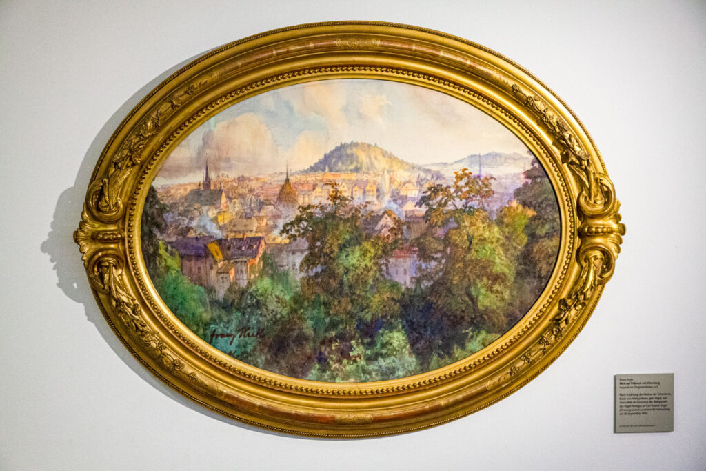 Aquarell-Gemälde im originalen ovalen Goldrahmen mit einer Stadtansicht von Pößneck, welche von Bäumen im Vordergrund und von den Hügeln der Altenburg im Hintergrund umgeben ist. Das Gemälde stammt von dem Pößnecker Ehrenbürger Prof. Franz Huth.