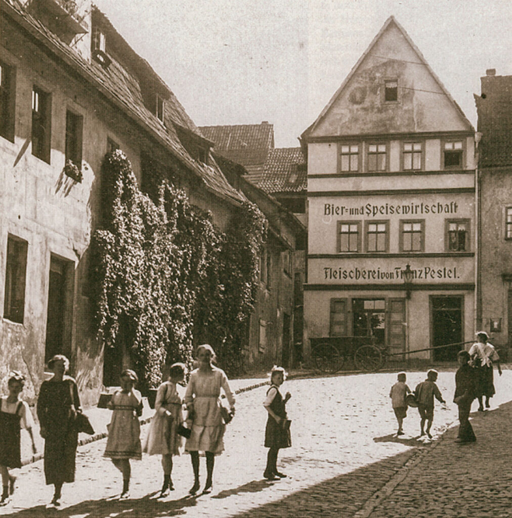 Historisches Gebäude, Klosterplatz 6, mit Beschriftung 'Bier- und Speisewirtschaft' und 'Fleischerei von Franz Pestel', davor spielende Kinder auf Kopfsteinpflaster. Links ein Haus welches mit Efeu bewachsen ist.