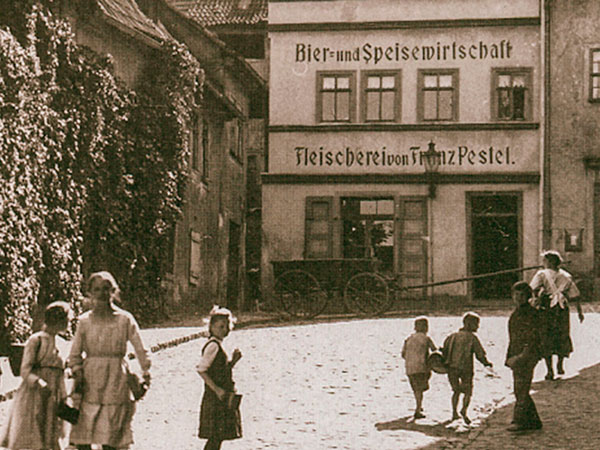 Historisches Gebäude, Klosterplatz 6, mit Beschriftung 'Bier- und Speisewirtschaft' und 'Fleischerei von Franz Pestel', davor spielende Kinder auf Kopfsteinpflaster. Links ein Haus welches mit Efeu bewachsen ist.