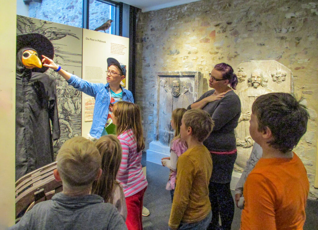 Acht Kinder und eine erwachsene Frau stehen vor der Figur eines „Pestarztes“, der in schwarz gekleidet ist und eine gelbe Schnabelmaske trägt. Rechts ein Teil der ehemaligen steinernen Stadtmauer mit Epitaphen.
