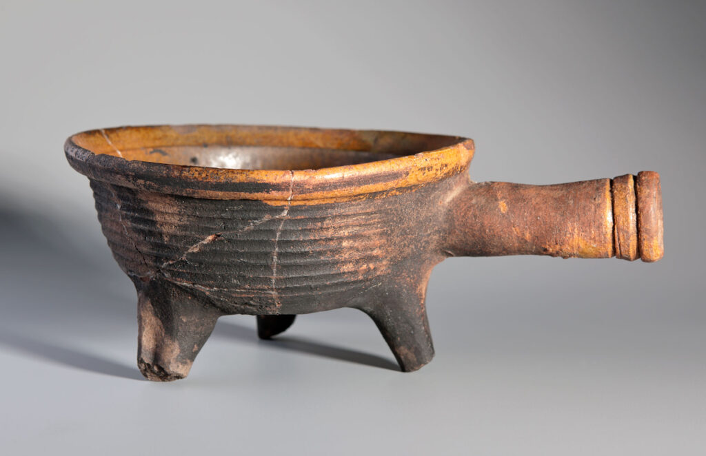 Eine dreifüßige, antike Tonschale mit einem Henkel. Die Schale ist dunkelbraun bis schwarz gebrannt und hat am oberen Rand einen ockerfarbenen Streifen. Der Henkel ist zylindrisch geformt und ebenfalls ockerfarben. Die Schale weist Gebrauchsspuren und kleine Beschädigungen auf.