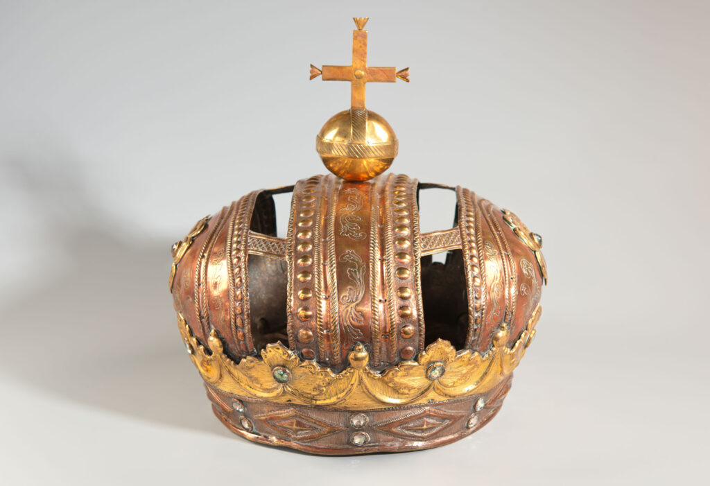 Eine detailreiche, runde Krone aus Kupfer- und Messingblech, welche aus mehreren übereinanderliegenden Bändern besteht, die mit floralen Mustern und kleinen Steinen verziert sind. Auf der Krone befindet sich ein Reichsapfel.